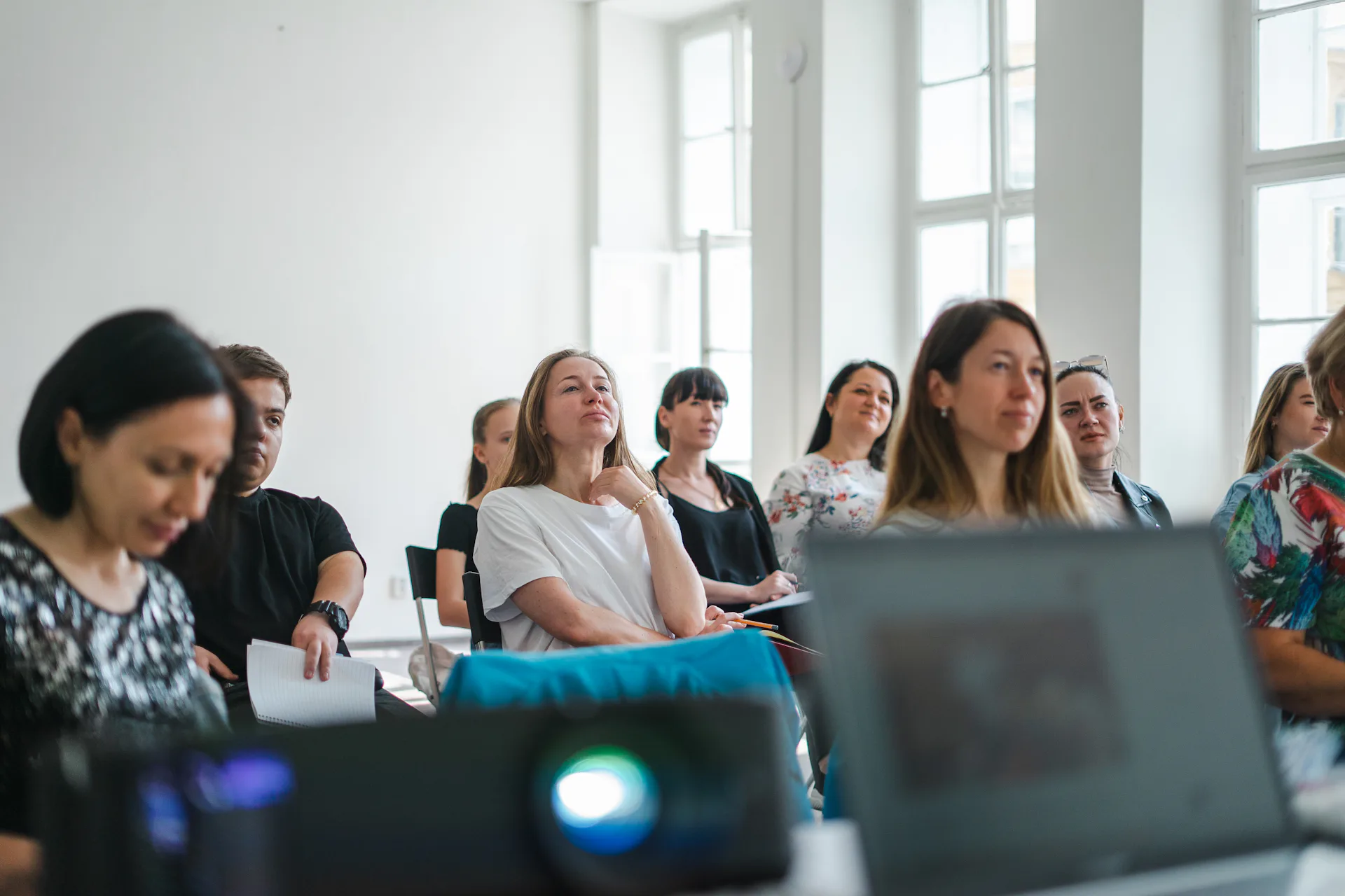 Adults seated in a sunlit room with tall windows, listening closely during a workshop